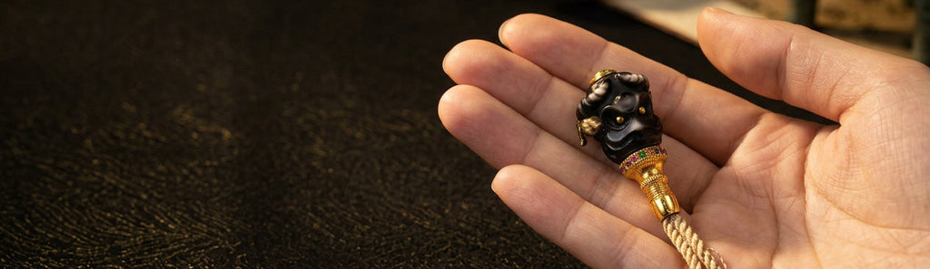 A close-up of a hand holding a decorative black and gold tassel bead pendant with a carved demon face and small multicolor gem accents.